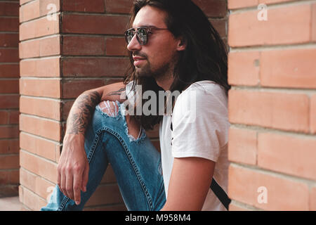 Attraente giovane uomo che indossa gli occhiali da sole e capelli lunghi rilassante in background urbano. Occhiali da sole modello maschile con i capelli lunghi e tatuaggi a portata di mano. Foto Stock