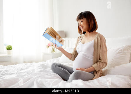 Felice incinta donna asiatica libro di lettura a casa Foto Stock