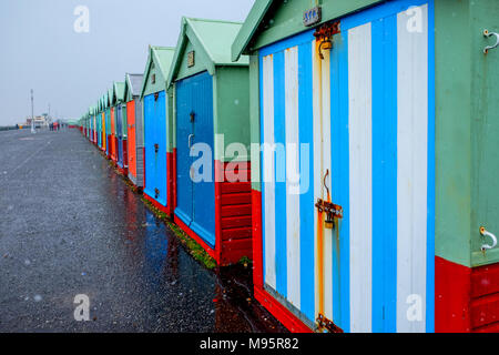 Brighton Seafront quaranta beach capanne, le capanne sono variopinte porte in una linea retta su un calcestruzzo promenade il cielo grigio è la spiaggia più vicina h Foto Stock