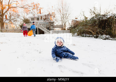 Ragazza sorridente seduto nella neve con altri bambini in corso di riproduzione in background Foto Stock