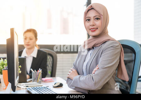 Imprenditrice musulmana di sorridere alla telecamera con fiducia e con il suo braccio mani incrociate. Si siede accanto al suo collaboratore in ufficio. Foto Stock