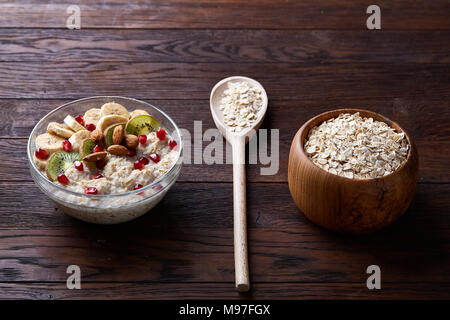 Composizione con vaschetta trasparente di fiocchi d'avena porrige con banana, kiwi, mandorla, pomergranate,asciugare i fiocchi d'avena in ciotola di legno e il cucchiaio di legno su vintage ba Foto Stock
