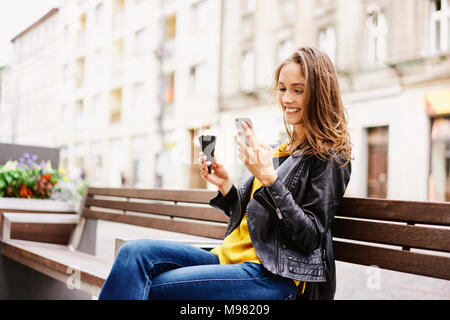 Ritratto di donna felice seduta sul banco di lavoro con cono gelato guardando al telefono cellulare Foto Stock