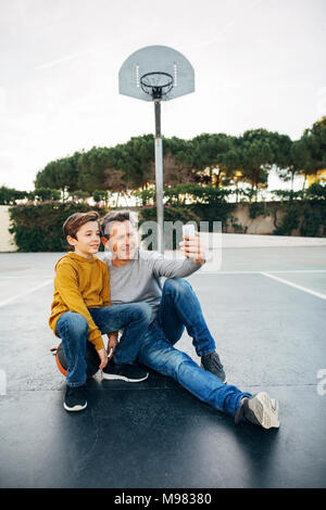 Padre e figlio seduti sul basket tribunale aperto prendendo un selfie Foto Stock