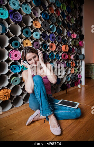 Rilassata donna matura ascoltando la musica di fronte all'assortimento di tappeti di yoga Foto Stock