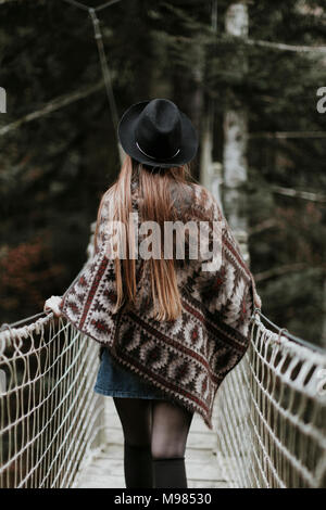 Vista posteriore del quartiere alla moda di giovane donna che indossa un cappello e il poncho camminando sul ponte di sospensione Foto Stock