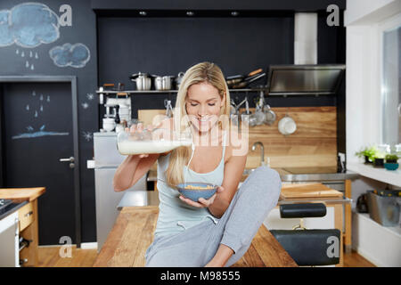 Donna bionda seduta sul tavolo da cucina versando il latte sul settore dei cereali Foto Stock