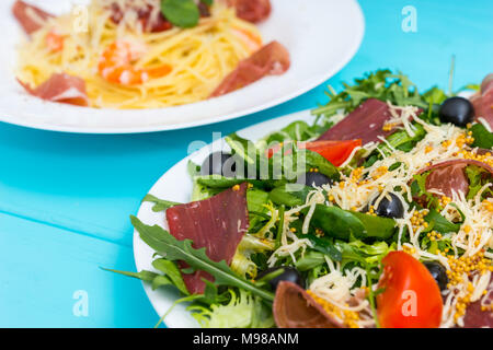 Close up di deliziosi con insalata di carne secca, prosciutto, formaggio grattugiato, le olive e il mix di foglie di lattuga in piastra bianca su blu tavolo in legno, spaghetti in Foto Stock