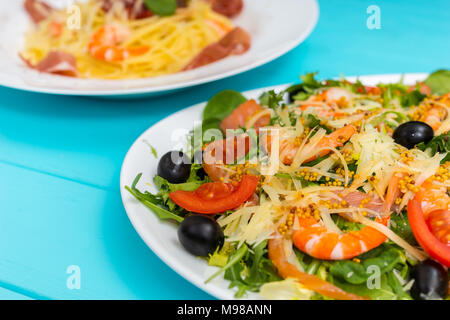 Close up di deliziosi con insalata di gamberetti, formaggio grattugiato, mix di foglie di lattuga, olive in piastra bianca su blu tavolo in legno, spaghetti in backgroun Foto Stock