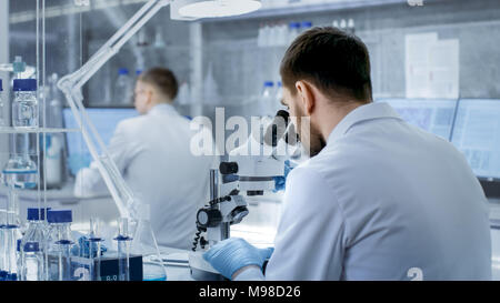 Colpo di Research Scientist regola il suo microscopio. Sta lavorando in una fascia alta laboratorio moderno con bicchieri, vetreria, microscopio e monitor Foto Stock