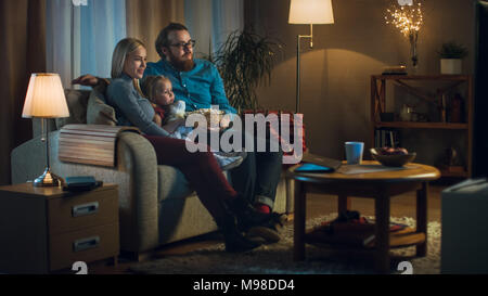 Colpo lungo di un padre, madre e bambina di guardare la TV. Essi sedersi su un divano nella loro accogliente soggiorno e mangiare popcorn. Si tratta di sera. Foto Stock