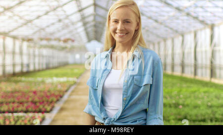 Colpo di bella bionda giardiniere sta sorridendo in una serra piena di fiori colorati. Foto Stock