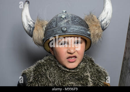 Viking brunette boy in pelliccia shirt in età prescolare in piedi in ambienti chiusi, indossando il costume hat con corna, in alto le mani, rendendo una faccia paura e spada Foto Stock