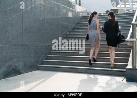 Due Asian office lady a piedi su per le scale e discutere gli uni con gli altri. In una vista posteriore. Sono entrambi di indossare tacchi alti e abiti formali dress borsa per il trasporto Foto Stock