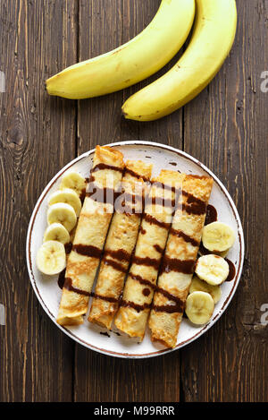 Delizioso rotolo di crêpe con fettine di banana e salsa al cioccolato su sfondo di legno. Vista superiore, laici piatta Foto Stock