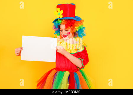 Bambina in costume da clown e hat holding tavolo bianco. Studio shot, isolato su sfondo giallo Foto Stock