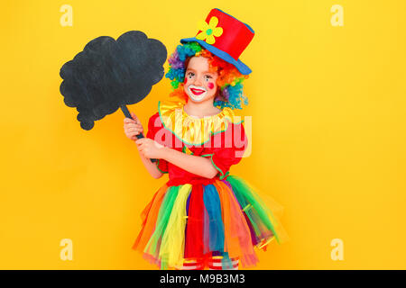 Bambina in costume da clown e cappello nero di contenimento tabella nella forma di una nuvola. Studio shot, isolato su sfondo giallo Foto Stock