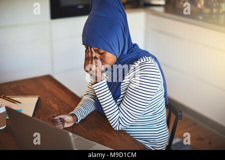 Ha sottolineato giovani arabi imprenditore femmina indossando un hijab seduta alla sua tabella di cucina usando un computer portatile mentre si lavora da casa Foto Stock