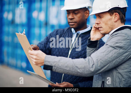 Gestore del porto in piedi con un collega in una spedizione commerciale cantiere parlando al cellulare e la lettura di un elenco inventario sulla clipboard Foto Stock