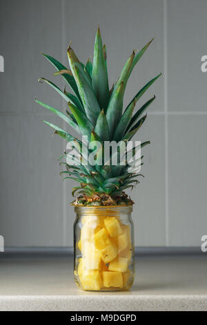 Fresh succoso e pezzi di ananas in un vasetto di vetro sul tavolo di cucina contro il grigio Sfondo rile Foto Stock