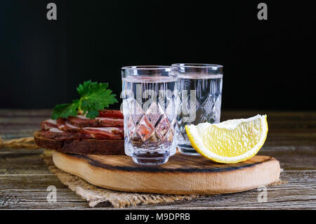 Due bicchierini di vodka con fettina di limone e pane di segale con Pancetta salata su sfondo scuro. Tradizionale bevanda forte. Close-up. Foto Stock