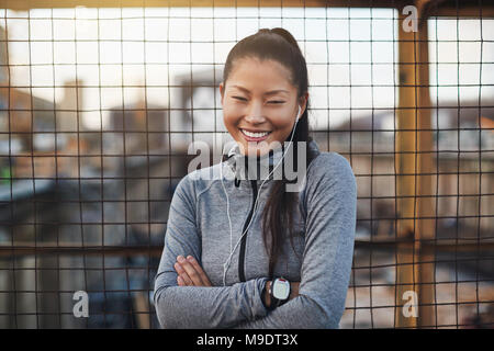 Giovane donna asiatica in esercizio marcia sorridente e indossare gli auricolari appoggiato contro una parete in mesh aperto con le braccia incrociate Foto Stock