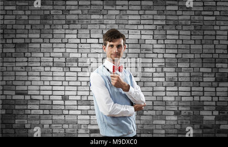 Inquadratura orizzontale di fiduciosa e giovane imprenditore della smart casual tubazione di fumare mentre permanente grigio contro un muro di mattoni su sfondo. Foto Stock