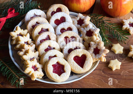 Linzer biscotti di Natale riempito con confettura di fragole e spolverizzati di zucchero, disposti su una piastra su un tavolo di legno, con mele e rami di abete rosso in t Foto Stock