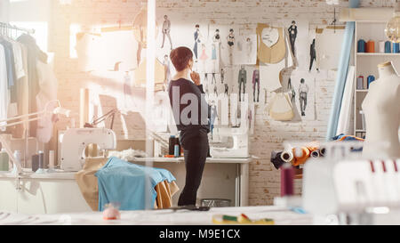Moda femminile, designer, guardando a disegni e schizzi che sono fissati alla parete dietro la sua scrivania. Studio è soleggiato. Personal Computer Foto Stock