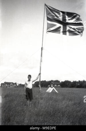 1934, storico, un giovane scout britannica in un campo erboso a un campo scout a Dublino, Irlanda, sorge poudly accanto ad alto pennone tenendo un Union Jack flag/ Foto Stock