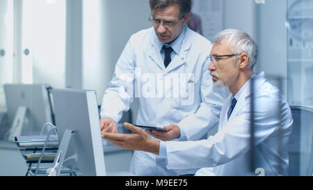 Due Senior Medici a discutere di problemi medici su computer desktop. Uno di loro Tenere computer Tablet. Office è moderno e luminoso. Foto Stock