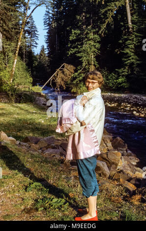 Giovane donna sorridente che indossa occhiali, pantaloncini e scarpe rosse che tengono un bambino addormentato avvolto in uno scialle vicino a un fiume, Spirit Lake, Washington state, USA negli anni '50 Foto Stock