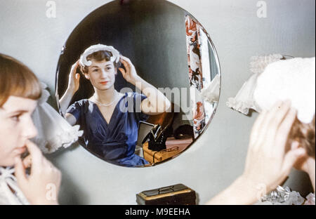 Giovane donna attraente che si prepara per la sua cerimonia di nozze mettendo su un cappello guardando in uno specchio. Indossa un abito da sposa blu scuro e una collana di perle mentre la sorella più giovane guarda Foto Stock