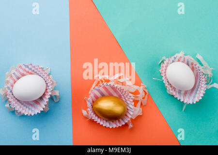 Tre uova di Pasqua in involucri di carta. Due bianchi e uno golden egg su carta colorata sfondo con copia spazio. Felice Pasqua concetto. Foto Stock