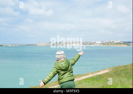 Retro del ragazzino in giacca calda sciarpa e hat steso le mani ai lati sul verde pendio contro il fondale di baia mare con barche a vela flottante in co Foto Stock