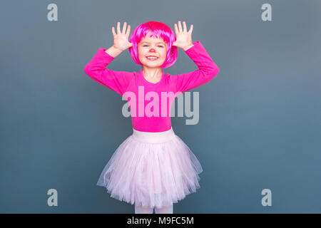 Ragazza in un carnevale rosa muta in una parrucca tiene le mani vicino alla sua testa. Studio shot, isolata su uno sfondo grigio Foto Stock