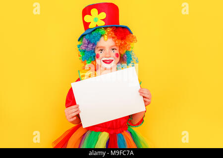 Carino bambina in costume da clown holding scrivania bianca per una copia dello spazio. Studio shot, isolato su sfondo giallo Foto Stock