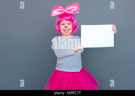 Attraente piccola ragazza in costume da clown holding scrivania bianca. Studio shot, isolata su uno sfondo grigio Foto Stock