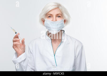 Le donne più anziane medico indossando maschera chirurgica studio isolato su sfondo bianco tenendo la siringa di anti-invecchiamento cercando di iniezione fotocamera sorridendo felice Foto Stock