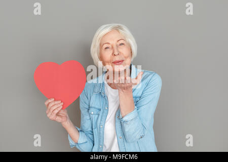 Le donne più anziane indossano jeans camicia studio isolato su sfondo grigio per celebrare il giorno di San Valentino di contenimento predisposto carta cuore di inviare aria kiss hap Foto Stock