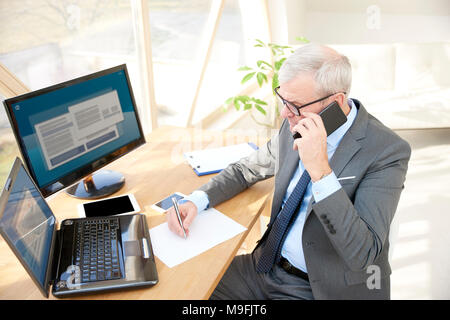 Alta angolazione del senior financial consultant imprenditore indossare tuta mentre seduto alla scrivania in ufficio nella parte anteriore del computer portatile ed effettuare una chiamata. Foto Stock