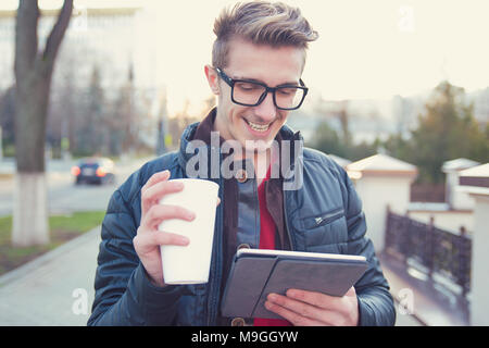 Giovane uomo sorridente in abbigliamento esterno e occhiali utilizzando tablet e di bere il caffè sulla strada. Foto Stock