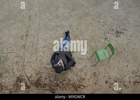 Uomo seduto su una sedia nel parco della città indossando cappotto e cappa, dormire, con le mani nelle tasche - vista aerea Foto Stock