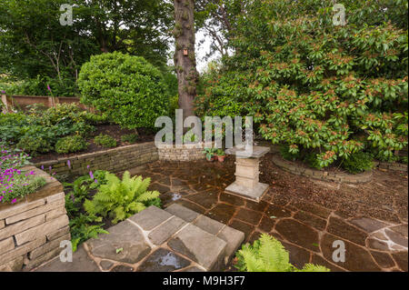 Piccolo pavimentato ombreggiata area terrazzata con pietra meridiana, bordo in rilievo, arbusti e piante - bella e tradizionale, giardino paesaggistico - Yorkshire, Inghilterra. Foto Stock