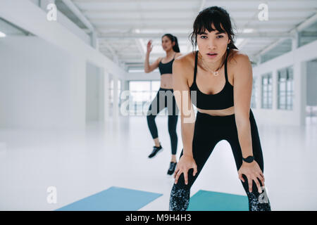 Sportive in piegato in avanti pone con le mani sulle ginocchia e amici di sesso femminile che esercitano in background. Donna prendendo break nella classe di esercizio in palestra. Foto Stock