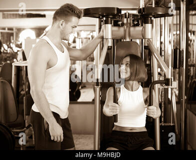 Ben addestrato uomo coach aiutare i giovani donna sorridente in utilizzando pec deck palestra macchine in ambienti chiusi Foto Stock