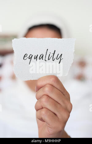 Primo piano di una giovane donna in interni indossando un bianco hijab che mostra un pezzo di carta nella parte anteriore del suo viso con la parità di testo scritto in esso Foto Stock