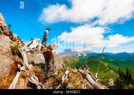 Un escursionista/fotografo guarda verso il basso dalla parte superiore della Cima del Castello, il suo cavalletto e fotocamera vicino con Mt. Sant Helens in background in Gifford Pinchot Na Foto Stock