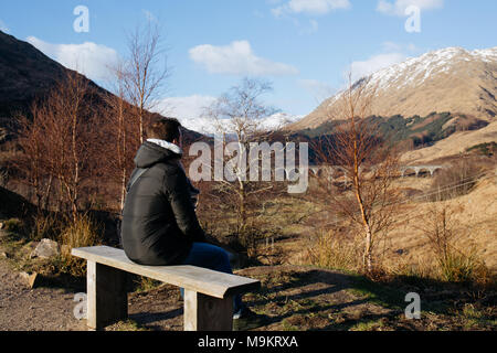 Un uomo si siede su una panca in legno, guardando il viadotto Glenfinnan, Scozia, su una soleggiata giornata di primavera. Foto Stock