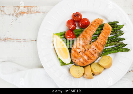 Fetta di salmone alla griglia guarnito con patate cotte, asparagi verdi, sauted pomodori ciliegia e limone su una tavola in legno rustico. Vista superiore Foto Stock